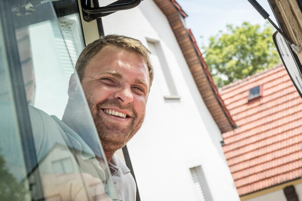 Freundlich lächelnder Mann lehnt sich aus einem Baufahrzeug vor einem Wohnhaus.